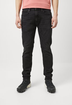 Mies, jolla on päällään kapeat mustat farkut ja mustat urheilukengät, seisoo yksinkertaisen valkoisen taustan edessä.