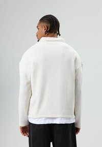 Long-sleeve, cream-colored sweater with a textured knit finish, highlighted by a collar and relaxed fit. Paired with black pants.