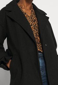Cappotto di lana nero con colletto a punte, tasche laterali e bottoni anteriori. Indossato sopra una camicia a stampa leopardata marrone e jeans.