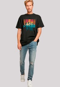 Zwarte t-shirt met een bergafbeelding in turquoise en oranje, gecombineerd met lichtblauwe distressed jeans en witte sneakers.