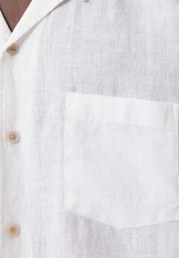 Camisa de lino blanca con un bolsillo en el lado izquierdo, detalles de botones y una tela texturizada y ligera.