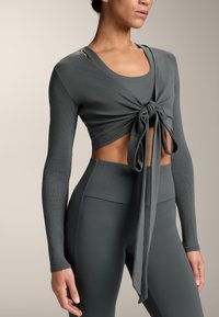 Sivi, dugih rukava crop top s čvorastim dizajnom sprijeda, uparen s tajicama visokog struka. Tkanina ima teksturirani, rebrasti finiš.