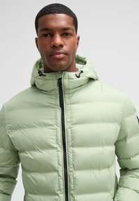 Giacca a vento color verde chiaro con design imbottito, cappuccio regolabile e zip nera. Presenta tasche laterali e un'etichetta del logo vicino al colletto.