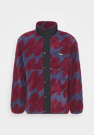 Chaqueta de forro polar con un patrón abstracto en rojo y azul, cierre frontal con botones a presión negros, cuello alto y logotipo de la marca en el pecho.