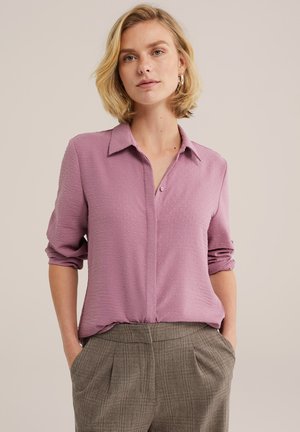 Femme aux cheveux blonds courts portant une chemise texturée mauve à boutons et un pantalon gris à carreaux, debout les mains dans les poches devant un fond neutre.