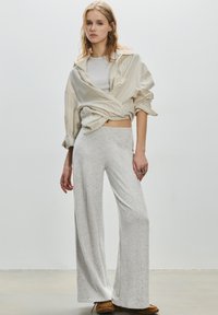 Chemise oversize légère à rayures beige, superposée sur un haut gris ajusté. Pantalon ample gris à jambes larges, texture lisse, associé à des chaussures marron.