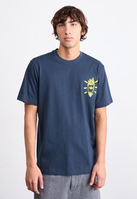 Ung man med vågigt brunt hår bär en marinblå T-shirt med en liten gul grafik av pommes frites i en kopp på bröstet, grå byxor.