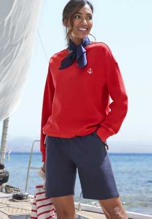 Red sweatshirt with anchor logo, paired with navy shorts. Model wears a blue scarf. Background features a seaside setting with a sailboat.