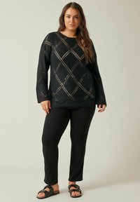 Schwarzer Strickpullover mit einem Rautenmuster, lockerer Passform und langen Ärmeln, kombiniert mit schwarzen Hosen und schwarzen Sandalen mit Schnallen.