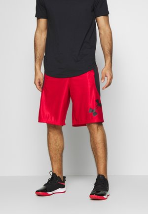 Hombre con camiseta negra, pantalones cortos deportivos rojos de Under Armour y zapatillas negras y rojas, de pie frente a un fondo liso.