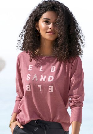 Pink long-sleeve top with a relaxed fit and white graphic text. Smooth fabric with ribbed cuffs. Paired with dark drawstring pants.