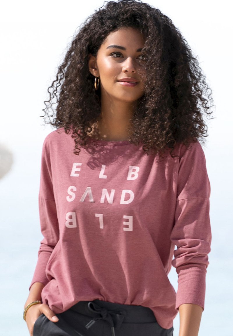 Pink long-sleeve top with a relaxed fit and white graphic text. Smooth fabric with ribbed cuffs. Paired with dark drawstring pants.