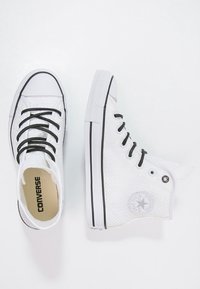 Bílé vysoké tenisky Converse s černými tkaničkami a gumovými podrážkami, jedna bota viděná shora, druhá ze strany.