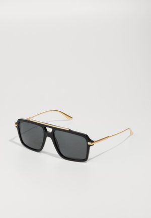 Gafas de sol negras en forma de cuadrado con acentos metálicos dorados, que cuentan con lentes oscuros y un marco grueso de plástico. Brazos delgados dorados se extienden desde el marco.