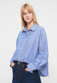 Πουκάμισο με κουμπιά σε μπλε και λευκές ρίγες, με άνετη εφαρμογή, διακοσμημένο με βολάν στο μπροστινό μέρος και μακριά μανίκια. Συνδυασμένο με σκούρο μπλε τζιν.