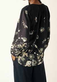 Schwarze Bluse mit floralen Mustern in grün und creme, mit gesmokten Schultern, langen Ärmeln und lockerer Passform. Kombiniert mit dunklen Hosen.