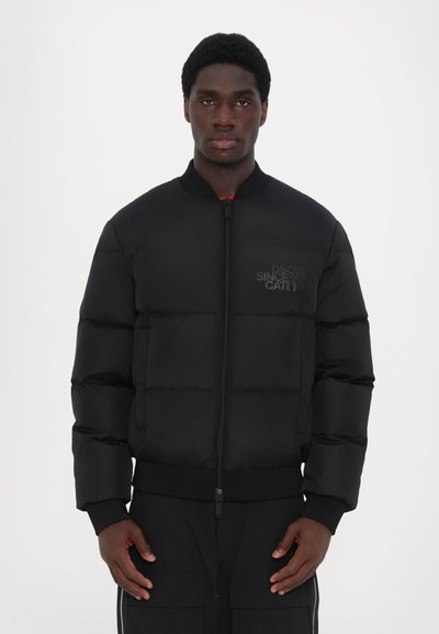 Doudoune noire avec fermeture éclair à l'avant, col et poignets côtelés. Présente un design matelassé et un détail de logo discret sur la poitrine gauche.