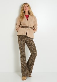 Femme portant une veste matelassée beige avec une ceinture à motif serpent, un haut rayé rouge, un pantalon à imprimé léopard et des chaussures pointues beiges, se tenant devant un mur blanc.