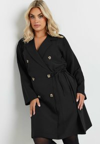 Cappotto nero doppiopetto con cintura in vita, realizzato in tessuto liscio. Presenta grandi bottoni dorati e maniche larghe.