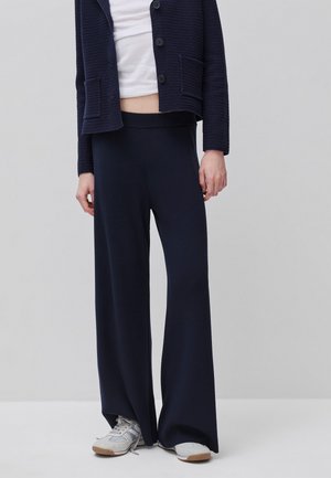 Osoba ubrana w granatowe szerokie spodnie, granatowy dziergany sweter z kieszeniami, biały krótki top i białe trampki na jednolitym szarym tle.