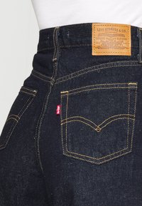 Dark blue denim jeans with yellow stitching, featuring a leather patch on the waistband and a red tag on the back pocket, tapered design.