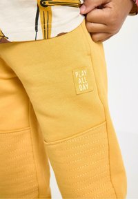 Gule træningsbukser med teksturerede paneler, der har et syet mønster og et lille mærke med teksten "PLAY ALL DAY." Blødt bomuldsmateriale.