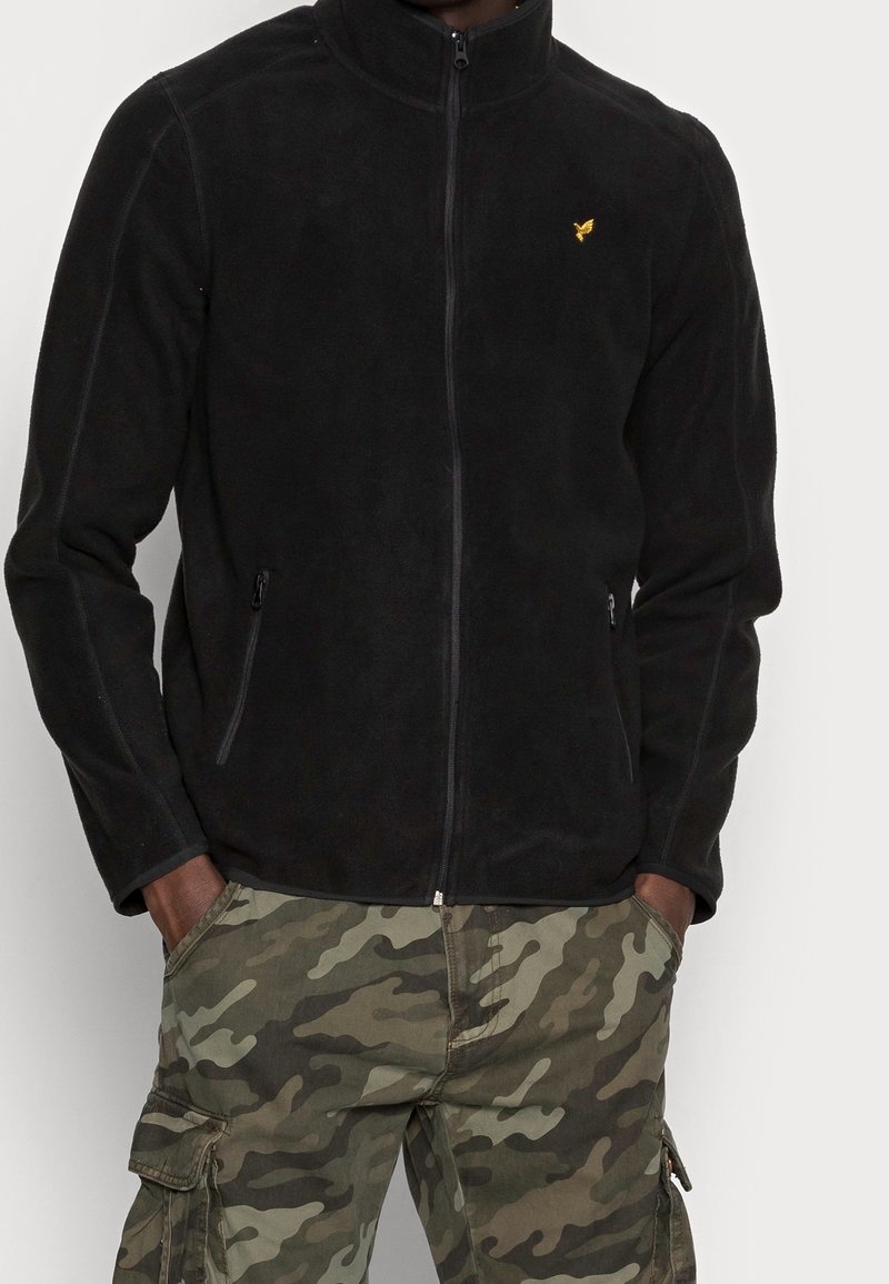 Homme portant une veste polaire noire zippée avec un logo d'oiseau jaune et un pantalon cargo camouflage, mains dans les poches, sur fond uni.