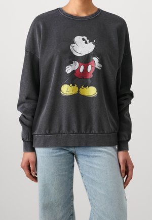 Osoba nosząca wyblakłą czarną bluzę z nadrukiem w stylu vintage z Myszką Miki w czerwonych szortach i żółtych butach, w połączeniu z jasnoniebieskimi dżinsami.