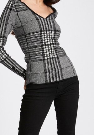 Pull à manches longues avec un col en V, présentant un motif en damier géométrique noir et blanc et un tissu texturé ; associé à un pantalon noir ajusté.