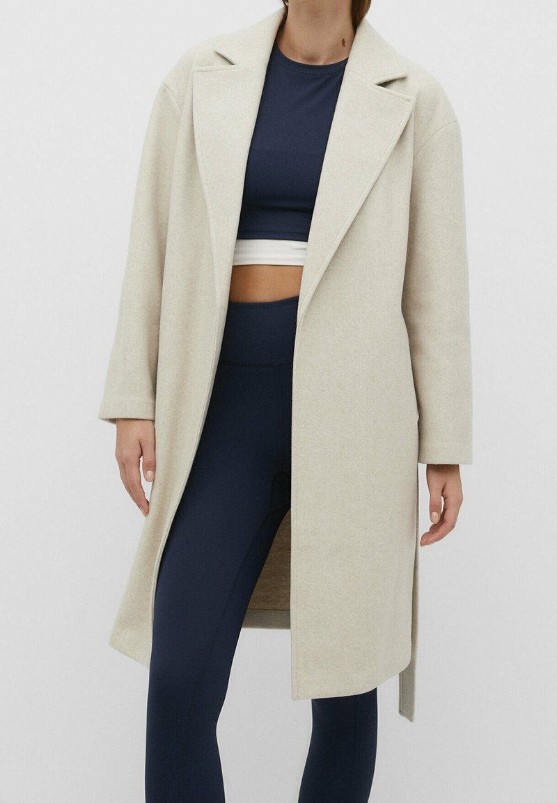 Femme portant un long manteau beige par-dessus un crop top bleu marine et des leggings bleu marine, debout devant un fond uni.