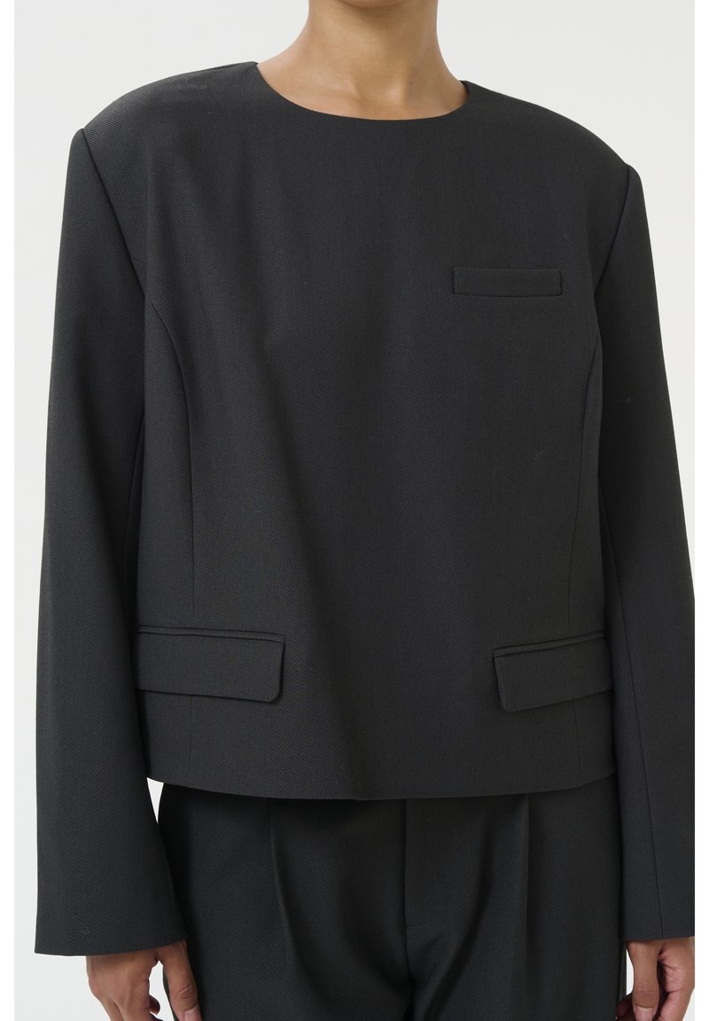 Blazer gris foncé cintré avec col rond et poches à rabat porté avec un pantalon assorti sur une personne visible des épaules aux hanches.