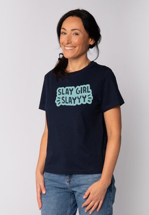 Vrouw met donker haar die glimlacht, gekleed in een marineblauw T-shirt met blauwe tekst "SLAY GIRL SLAYYY" en lichtblauwe spijkerbroek, tegen een effen achtergrond.