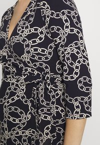 Robe marine à manches trois-quarts, présentant un motif de maillons en argent. Le tissu est doux avec un détail de taille nouée pour un look ajusté.