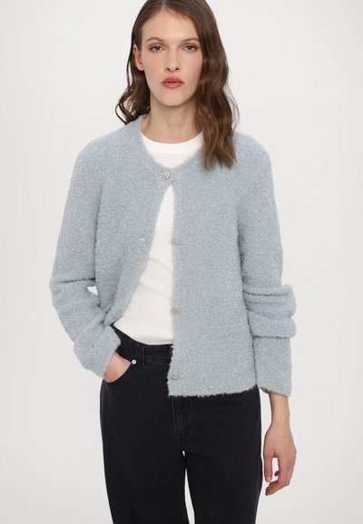 Blå, fluffy cardigan med lange ermer, rund halslinje og dekorative sølvknapper, iført over en hvit topp og svarte bukser.