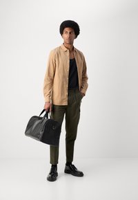 Bolsa de viaje de cuero negro, camisa de botón color marrón claro sobre una camiseta negra sin mangas, pantalones verde oliva y zapatos negros de cordones. Modelo de pie sobre un fondo liso.