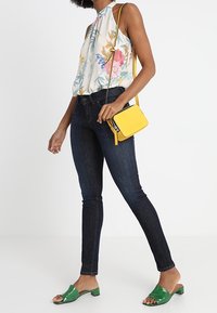 Blusa de halter floral en colores claros, jeans oscuros ajustados, sandalias planas verdes y un pequeño bolso amarillo tipo bandolera.