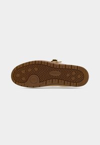 Semelle d'une sneaker Lacoste marron montrant un motif de grip en caoutchouc texturé et le nom de la marque, sur un fond blanc uni.