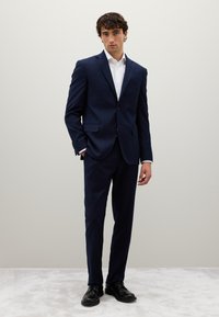 Completo a quadri blu navy realizzato in tessuto dalla texture liscia, con giacca monopetto e pantaloni slim fit, abbinati a scarpe nere.