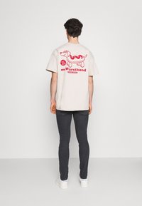 Camiseta beige con un gráfico juguetón de un dachshund con un diseño de perrito caliente en la parte posterior. Mangas cortas, corte relajado. Usada con jeans oscuros.
