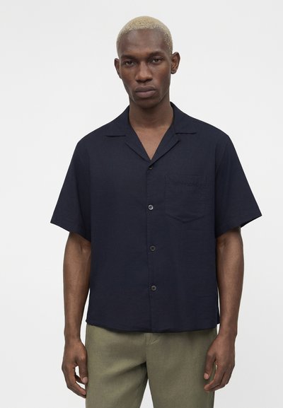 Νέος άνδρας με κοντά ξανθά μαλλιά που φοράει μαύρο κοντομάνικο πουκάμισο και ελιά πράσινο παντελόνι σε απλό λευκό φόντο.