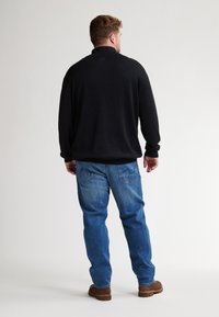 Schwarzer Pullover mit hohem Kragen, kombiniert mit hellblauen Jeans und braunen Schuhen, von hinten betrachtet.