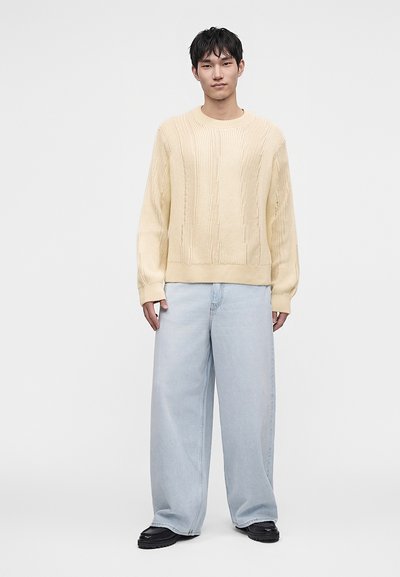 Jeune homme aux cheveux courts et noirs portant un pull en maille torsadée couleur crème, un jean large bleu clair et des chaussures noires, debout devant un fond uni.