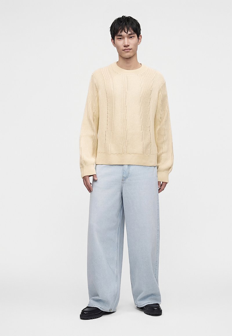 Young man with short black hair wearing a cream cable-knit sweater, light blue wide-leg jeans, and black shoes standing against a plain background.