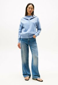 Helles blaues Zip-Hoodie, lässige Passform, gerippte Bündchen, kombiniert mit weit geschnittenen Jeans im hellen Waschung und schwarzen Flip-Flops. Schlichtes Design.