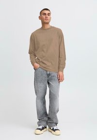 Langärmliges, hellbraunes Baumwollhemd mit Rundhalsausschnitt, kombiniert mit hellgrauen Jeans und schwarz-weißen Sneakern. Insgesamt ein einfaches Design.