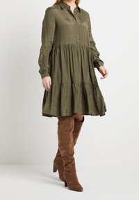 Olive green, długa sukienka koszulowa z warstwowym dołem, z guzikiem z przodu. Połączona z brązowymi zamszowymi botkami za kolano.