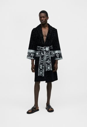 Άνδρας μοντέλο που φορά μαύρη και ασημένια ρόμπα μέχρι το γόνατο με ζώνη, όρθιος ξυπόλητος σε απλό λευκό φόντο.