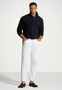 Fekete, hosszú ujjú galléros póló, fehér, szűk szabású farmernadrág és fekete mokaszinek. A mellkason egy kis piros logó található.