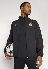 Sort sportjakke med lynlås krave, røde detaljer og Benfica-logo. Modellen holder en fodbold. En sort skjorte og bukser fuldender outfittet.