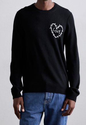 Hombre vistiendo un suéter negro de manga larga con un corazón de alambre de púas blanco y el texto "LOVE" en el pecho, combinado con jeans azules.